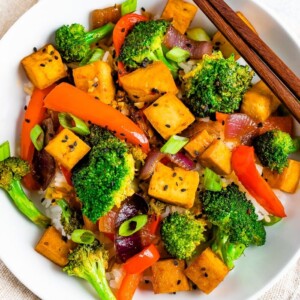 Bowl of rice with tofu and vegetable stir fry on top. Chopsticks are resting on the bowl.