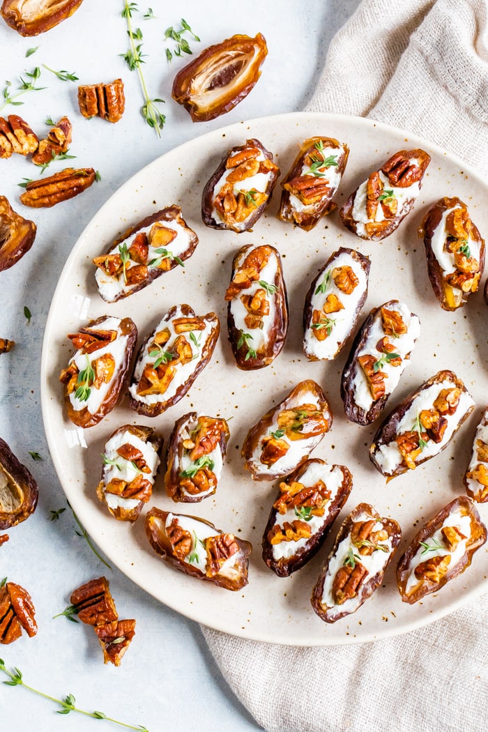 Plate with goat cheese stuffed dates topped with roasted pecans and fresh thyme. Ingredients in this easy recipe are around the plate along with a cloth napkin.