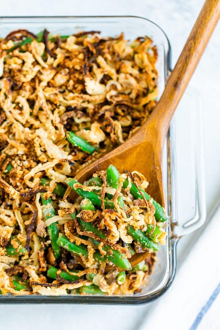 Casserole dish with healthy green bean casserole topped with crispy onions. A wood spoon is scooping some of the casserole.