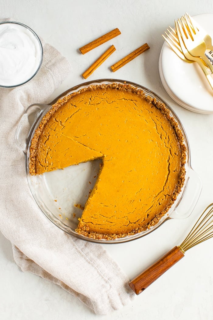 Vegan pumpkin pie with a slice taken out of it. The pie is on a table with plates, forks, and cinnamon sticks.