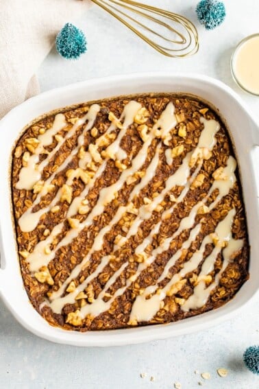 Gingerbread baked oatmeal drizzled with frosting. Baking dish is surrounded but a bowl of icing, mini Christmas trees, and a whisk.