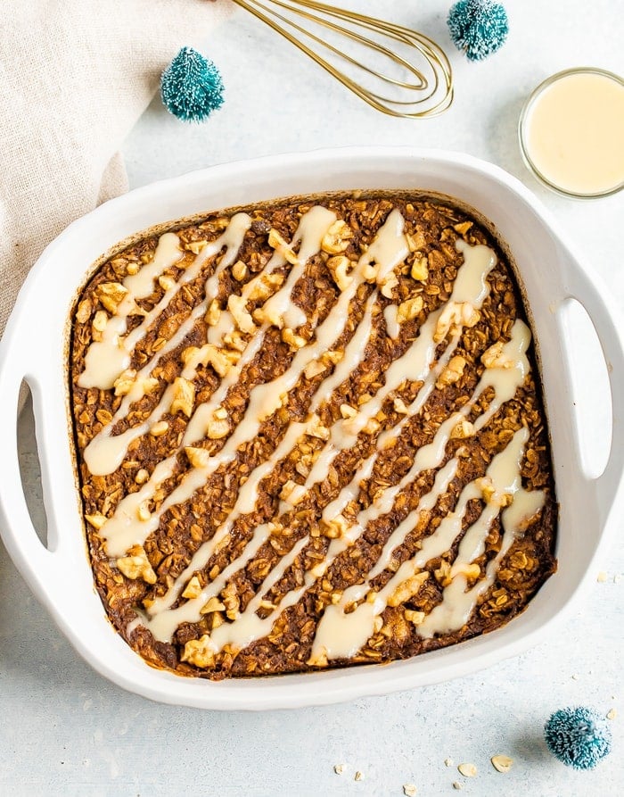 Gingerbread baked oatmeal drizzled with frosting. Baking dish is surrounded but a bowl of icing, mini Christmas trees, and a whisk.