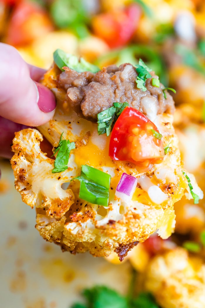 Hand holding a "cauliflower nacho" that's topped with cheese, beans, tomato and onion.