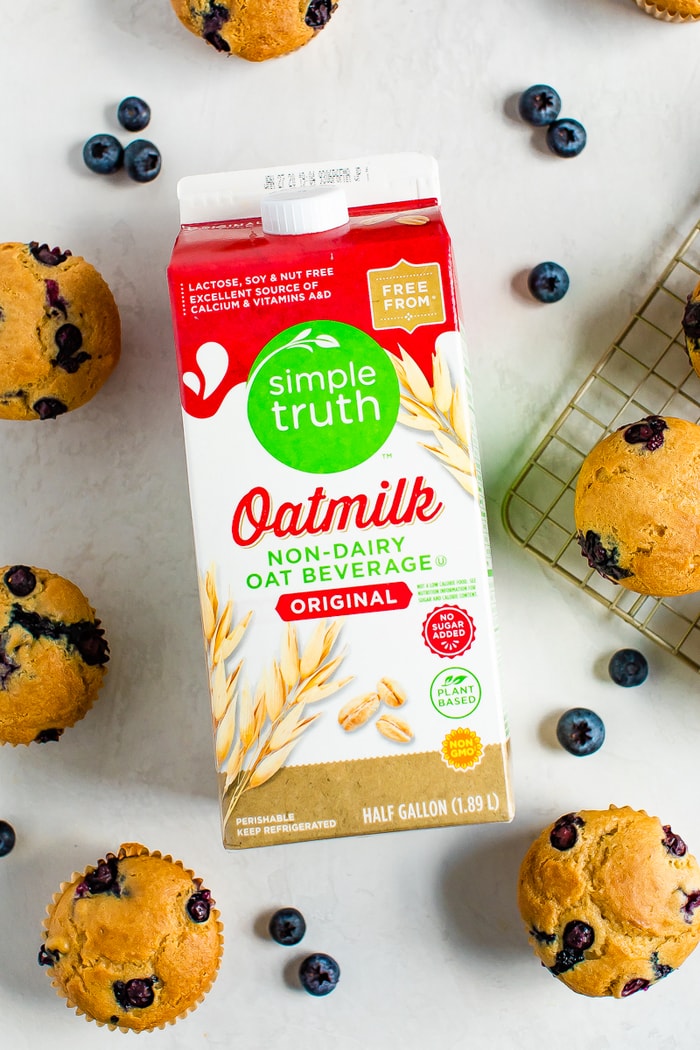 Carton of oatmilk on a table, surrounded by blueberry muffins and blueberries.