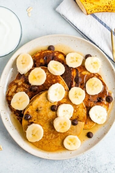Four Greek yogurt pancakes on a plate topped with bananas, chocolate chips and maple syrup.