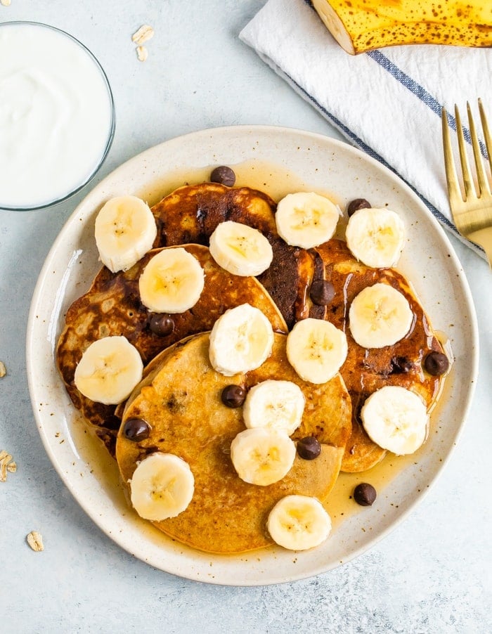 Four Greek yogurt pancakes on a plate topped with bananas, chocolate chips and maple syrup.