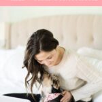 Woman working on a laptop while sitting on her bed with her small dog.
