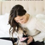 Woman working on her laptop while sitting on her bed with her dog.