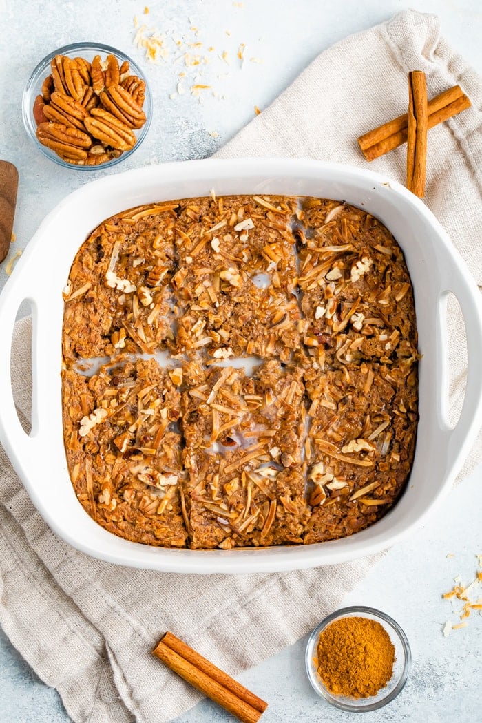 Square baking dish with chai baked oatmeal. Dish is on a table surrounded by a bowl of pecans, ground cinnamon and cinnamon sticks.