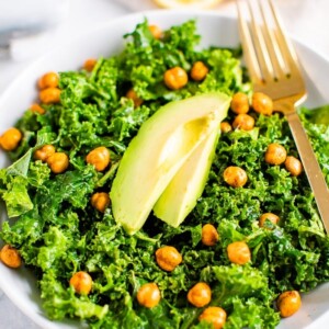 Bowl with kale and topped with lemon and avocado slices and a fork. Lemon halves on the side.