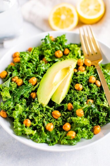 Bowl with kale and topped with lemon and avocado slices and a fork. Lemon halves on the side.