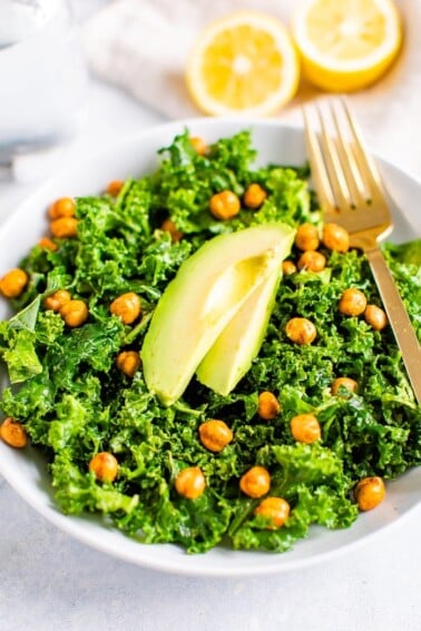 Bowl with kale and topped with lemon and avocado slices and a fork. Lemon halves on the side.