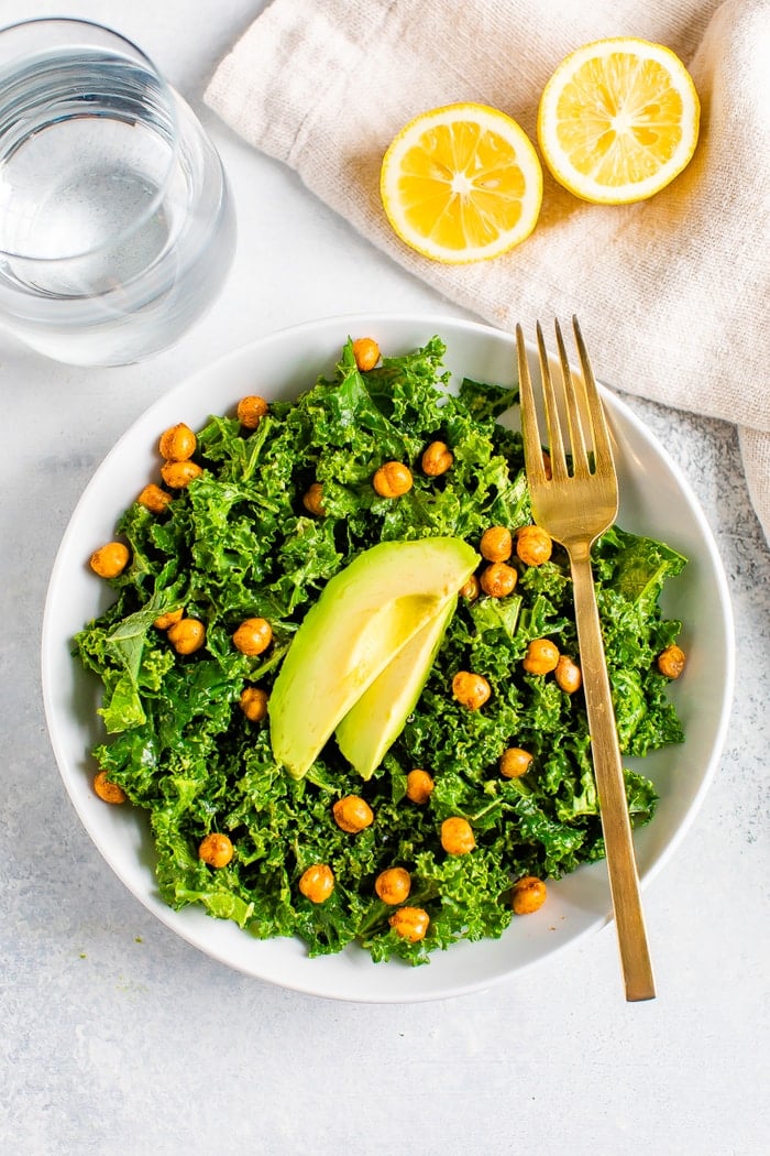 Kale salad topped with chickpeas and avocado. A fork is resting on the bowl, and slices of lemons and glass of water are next to the bowl.