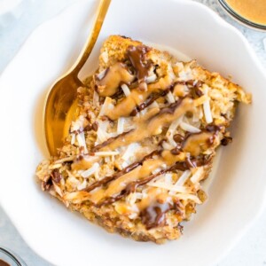 Slice of samoa baked oatmeal in a bowl with a gold spoon. Oatmeal is topped with coconut flakes and drizzles of caramel and chocolate.