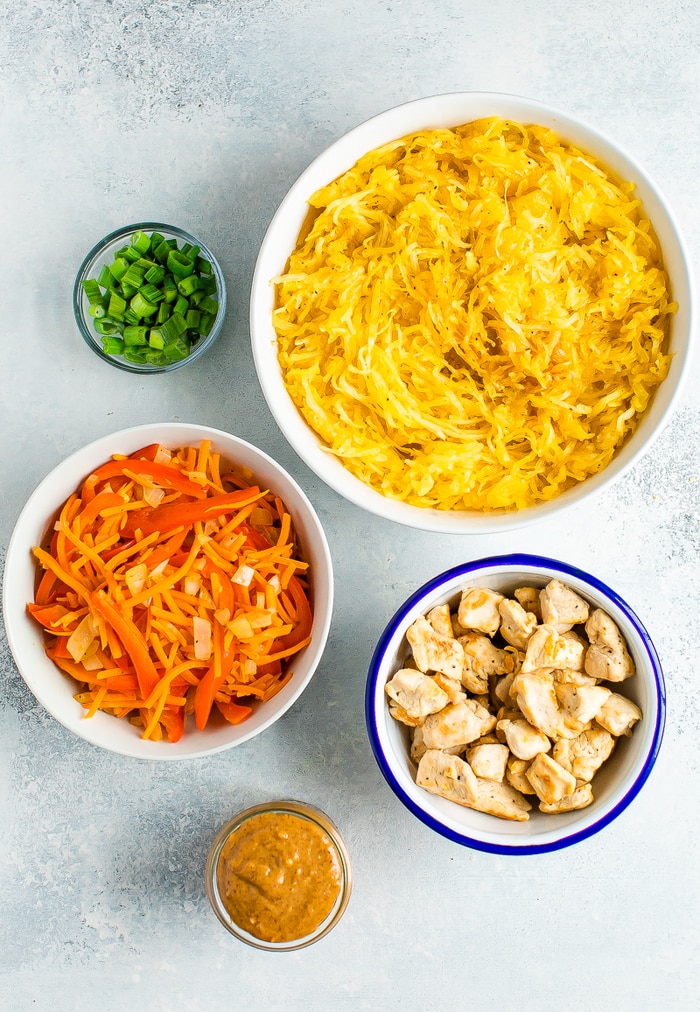 Ingredients to make spaghetti squash pad thai in bowls: green onions, spaghetti squash, carrots and peppers, chicken and almond butter.