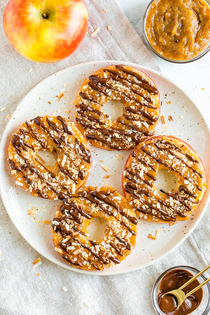 Four apple samoas on a plate. An apple, bowl of date caramel and a bowl and measuring spoon of chocolate are around the plate on the table.