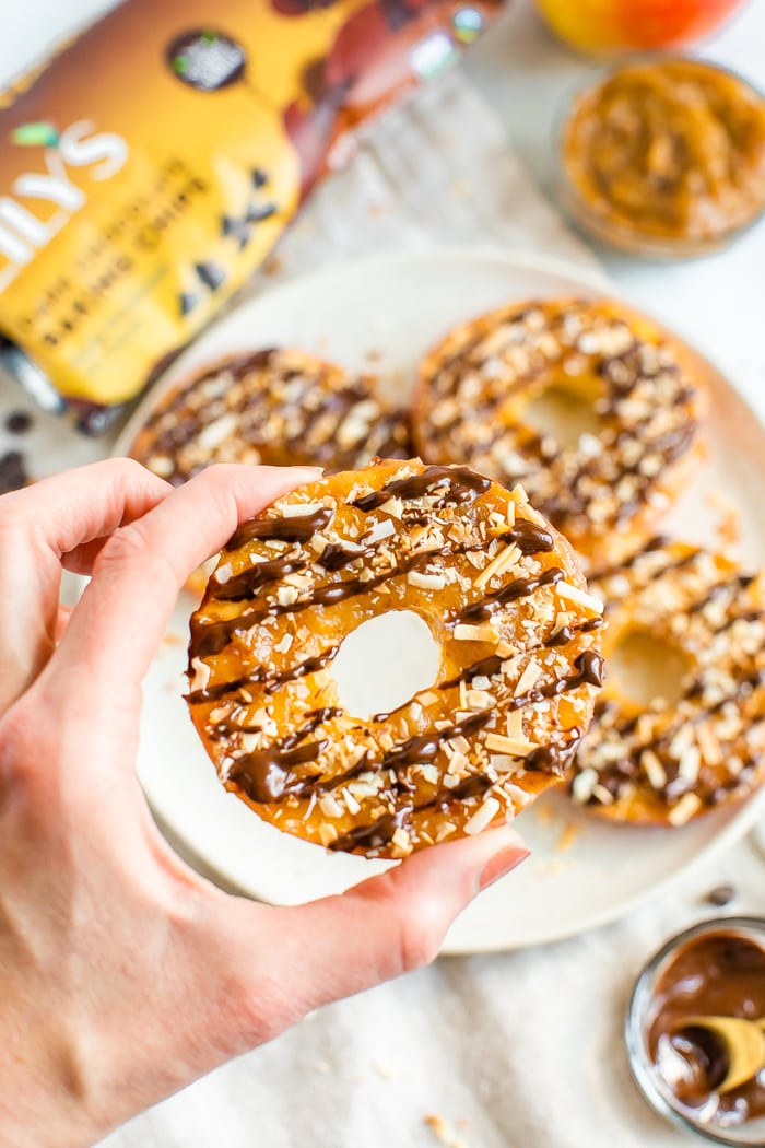 Hand holding an apple samoa which is an apple ring with date caramel, drizzled chocolate and toasted coconut.