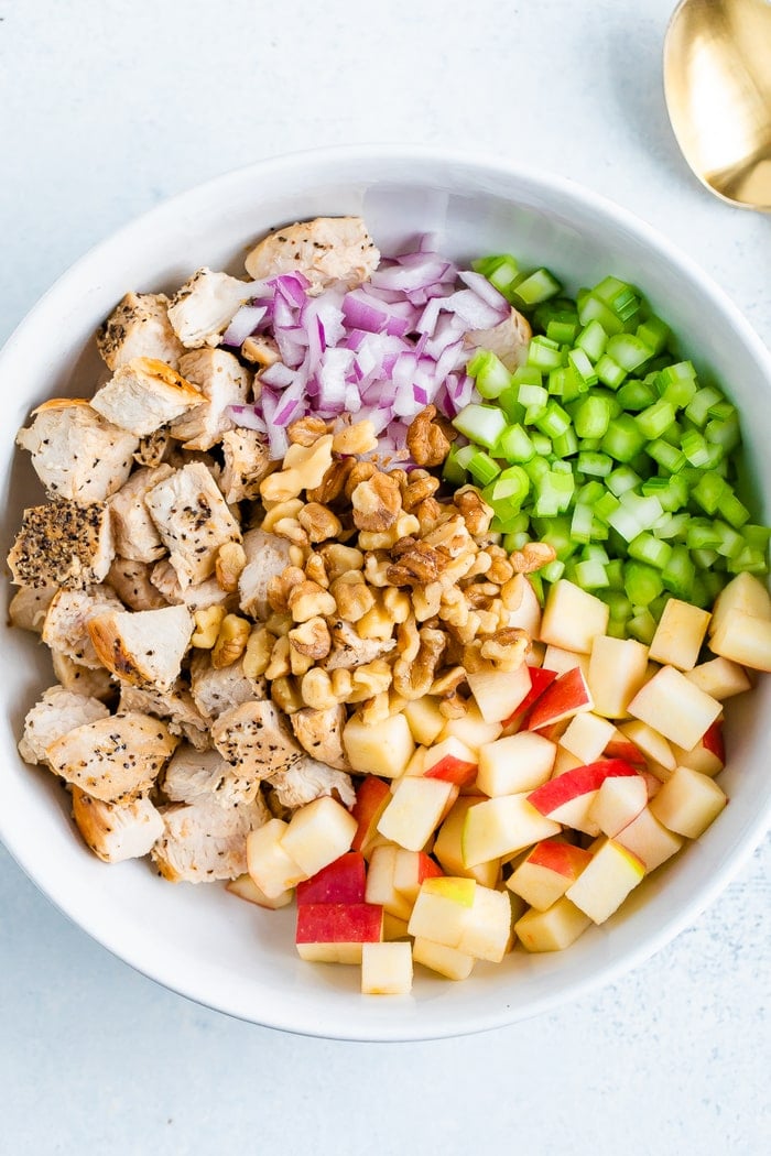 Bowl with the ingredients for a chicken waldorf salad before being mixed-- chopped grilled chicken, red onion, celery, walnuts and apples.