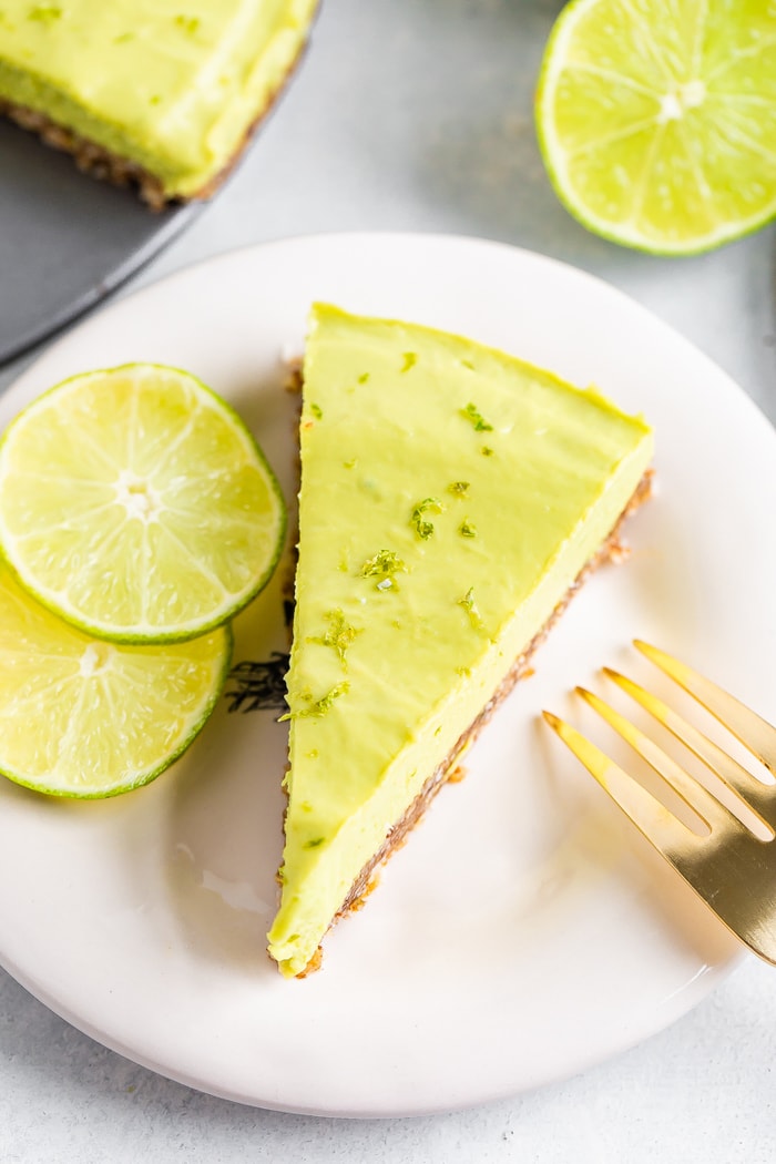 Slice of avocado lime tart on a plate with a gold fork and two slices of lime.