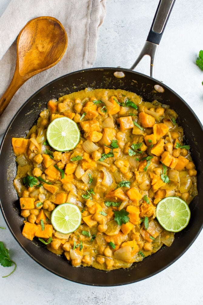 Skillet with curried sweet potatoes, onions and chickpeas in a coconut sauce. Topped with lime and cilantro. A wood spoon is beside the skillet.