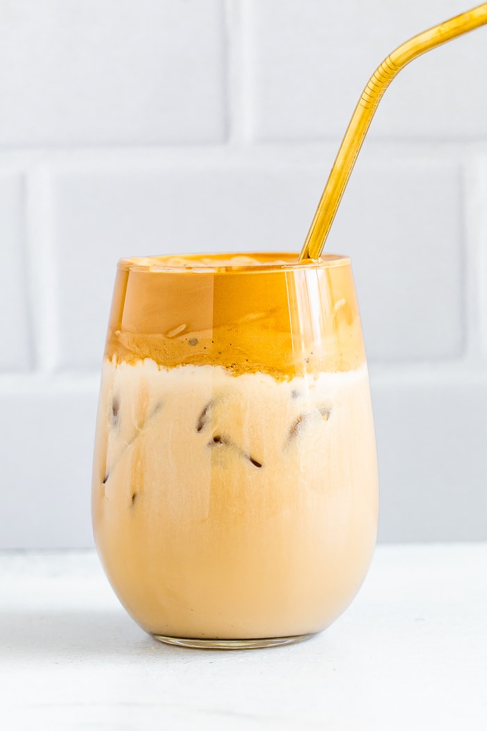 Glass of whipped coffee/dalgona coffee. The whipped coffee on top has been mixed into the almond milk and ice. A gold straw is in the glass.