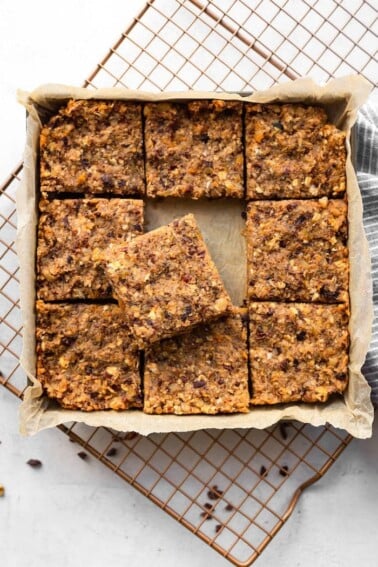 Square pan with fruit and nut bars cut into 9 slices. The middle square is sitting on top of the surrounding square pieces. The pan is sitting on a cooling rack.