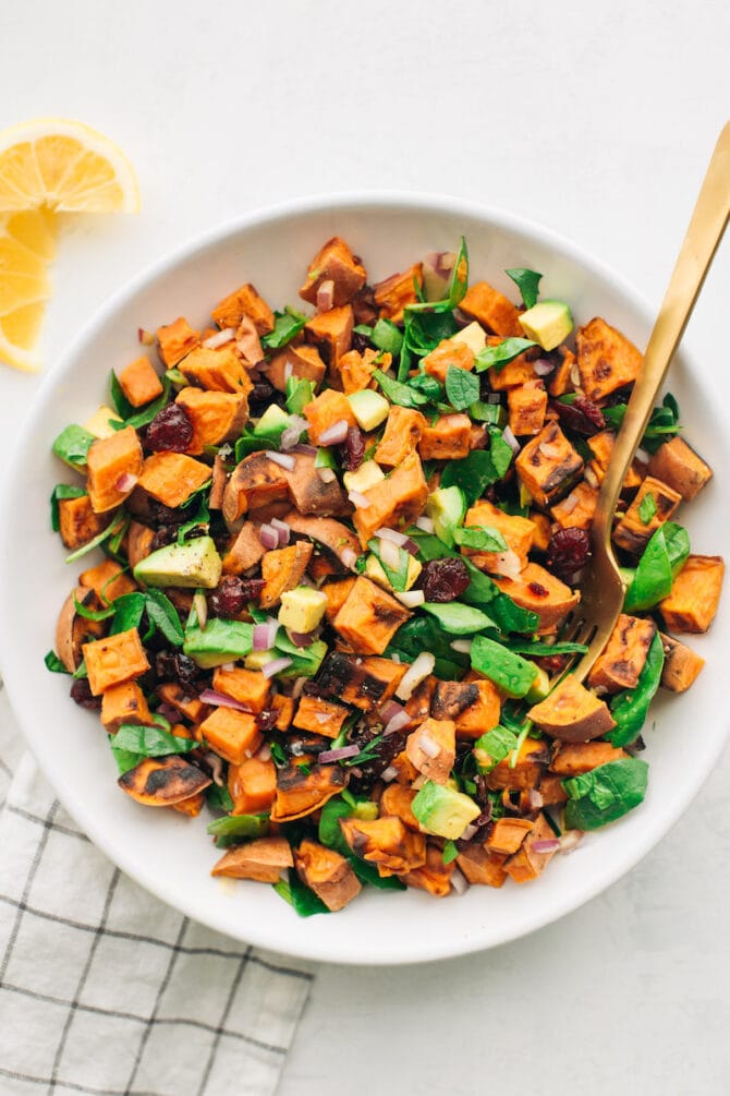 Roasted sweet potato salad with spinach, avocado, red onion and dried cranberries in a white bowl with a gold fork..