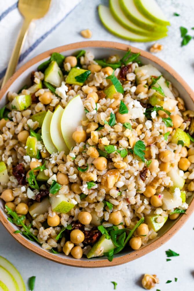 Bowl of barley salad with chickpeas and pear. Table is scattered with herbs and walnuts.