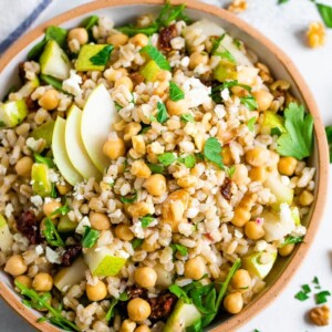 Bowl of barley salad with chickpeas and pear. Table is scattered with herbs and walnuts.