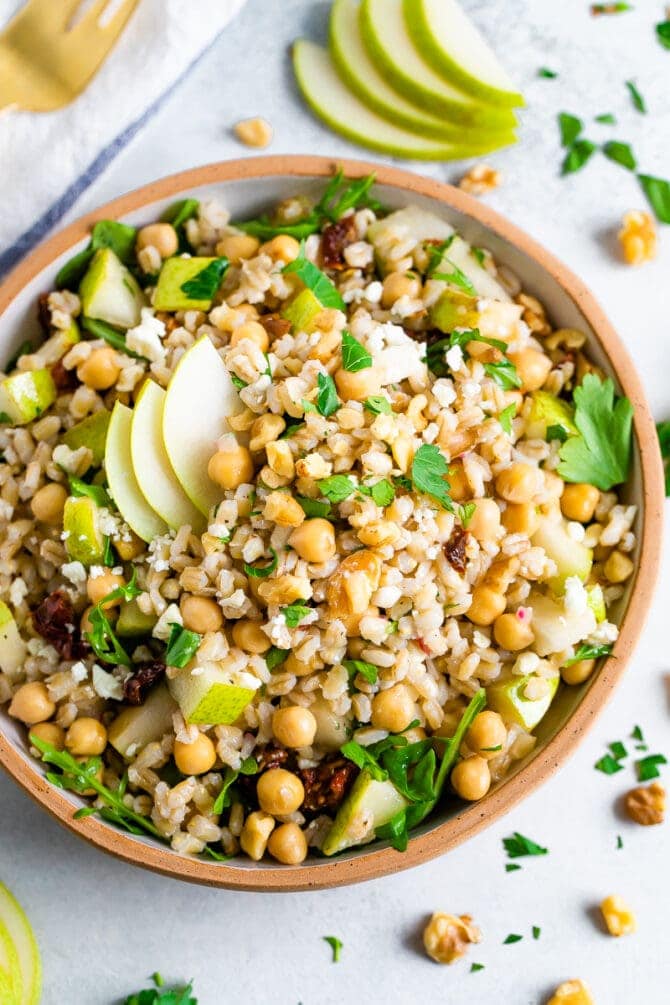 Bowl of barley salad with chickpeas and pear. Table is scattered with herbs and walnuts.