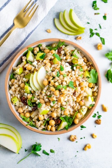 Bowl of barley salad with chickpeas and pear. Table is scattered with herbs and walnuts.