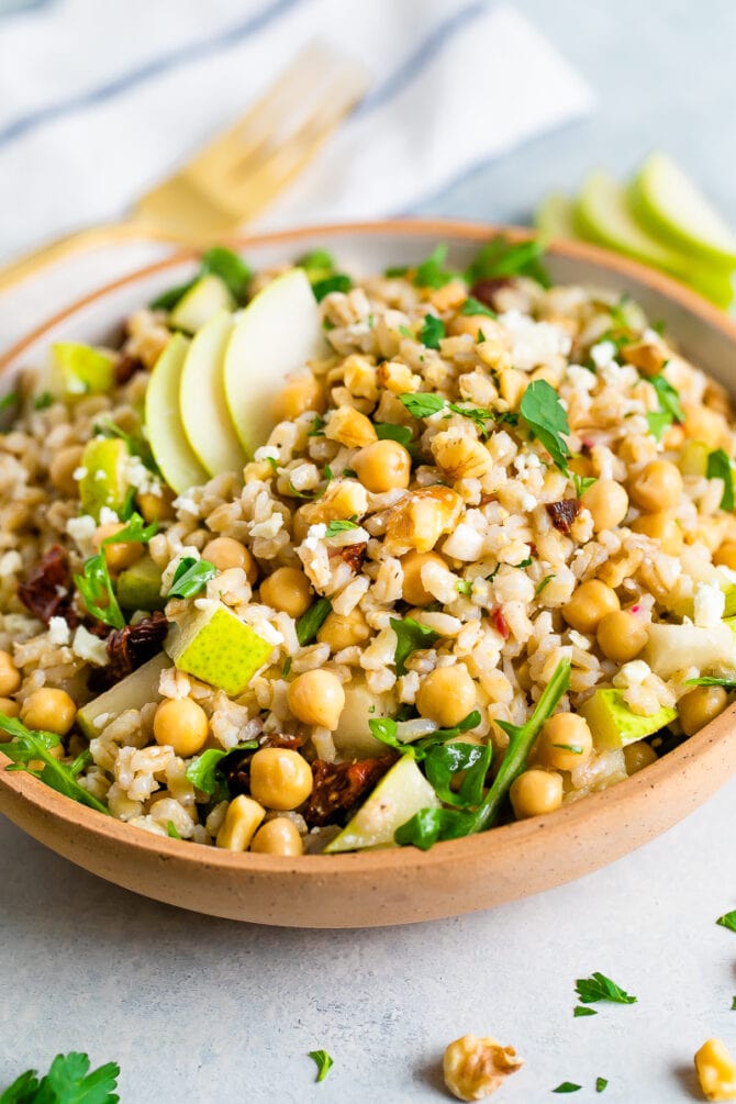 Bowl of barley salad with chickpeas and pear. Table is scattered with herbs and walnuts.