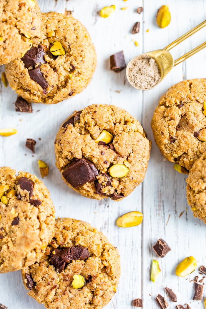 Dark chocolate chunk cookies with pistachios on a white table. A measuring spoon with cardamom and chocolate chunks and pistachios are around the cookies.