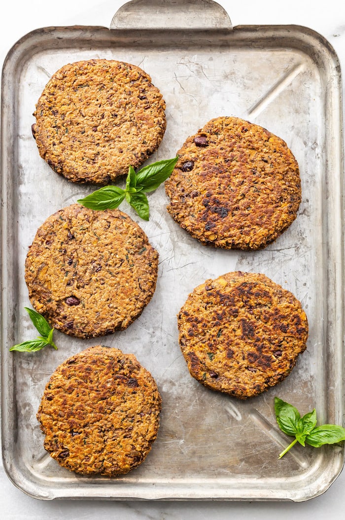 Five black bean sweet potato burgers on a baking sheet surrounded by basil leaves.