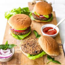 Three black bean sweet potato burgers on buns on a wood cutting board. A bowl of ketchup and onions are beside the burgers.