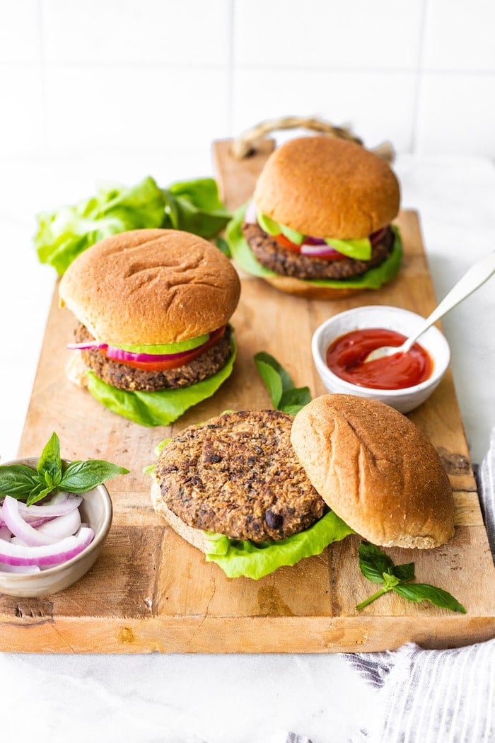 Three black bean sweet potato burgers on buns on a wood cutting board. A bowl of ketchup and onions are beside the burgers.