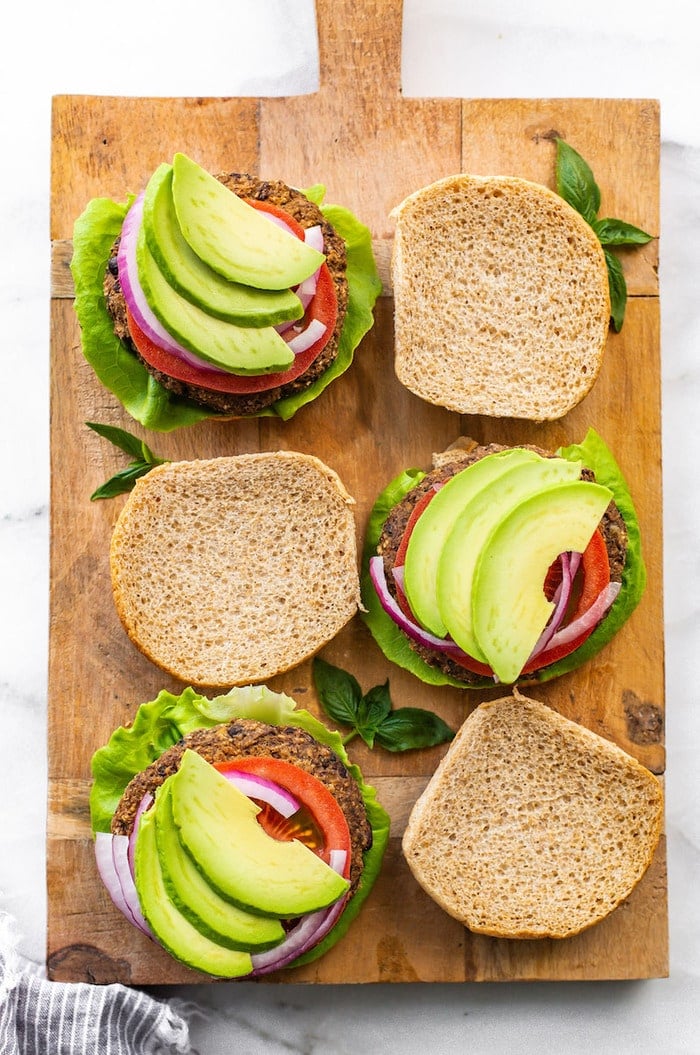 Three sweet potato black bean burgers on a wood board with the top bun off. Burgers are topped with tomatoes, onion, lettuce and avocado,