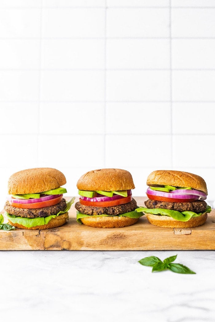 Three black bean sweet potato burgers on a wood cutting board. Burgers are on buns with lettuce, tomato, onion and avocado.