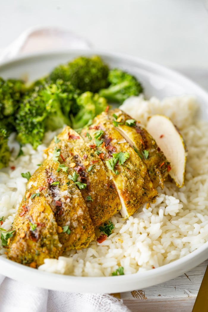 Sliced curry rub chicken breast topped with cilantro and red pepper flakes. Chicken is served over a bed of rice and next to broccoli.