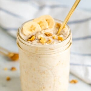 Mason jar with banana bread overnight oats, topped with banana slices and walnuts. Mason jar has a gold spoon handle sticking out.