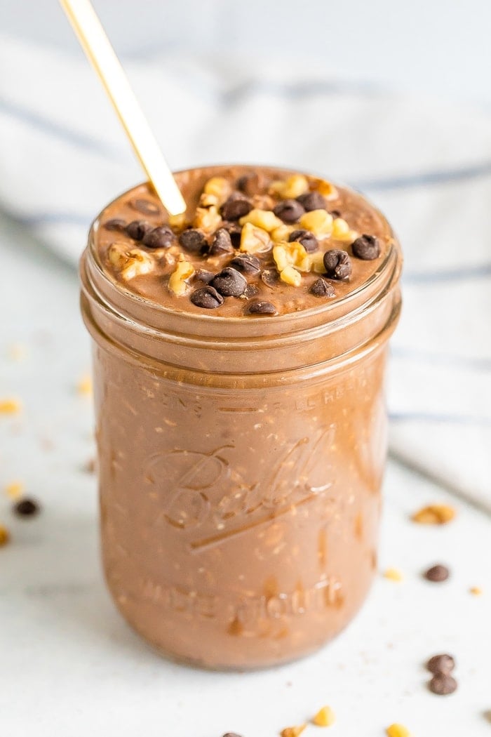 Chocolate brownie batter overnight oats in a mason jar with a gold spoon. Topped with walnuts and chocolate chips.
