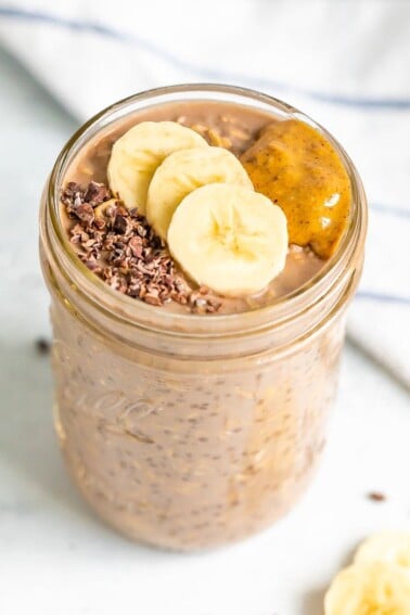 A mason jar with chocolate banana overnight oats topped with banana slices, cacao nibs and almond butter.