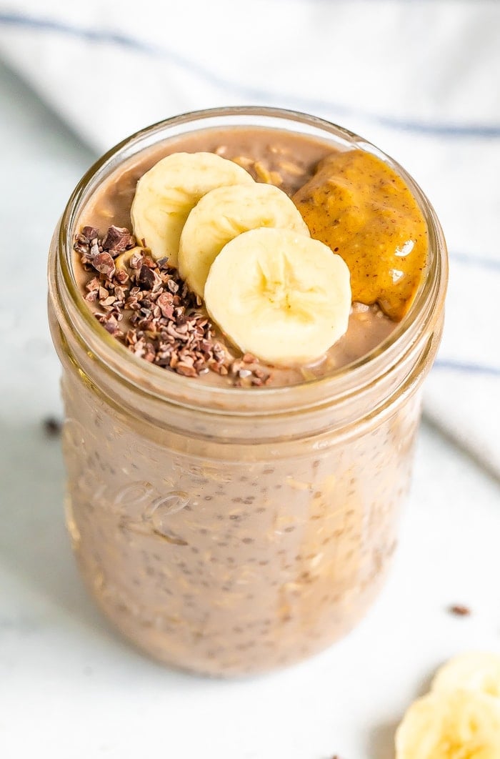 A mason jar with chocolate banana overnight oats topped with banana slices, cacao nibs and almond butter.