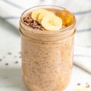 A mason jar with chocolate banana overnight oats topped with banana slices, cacao nibs and almond butter.