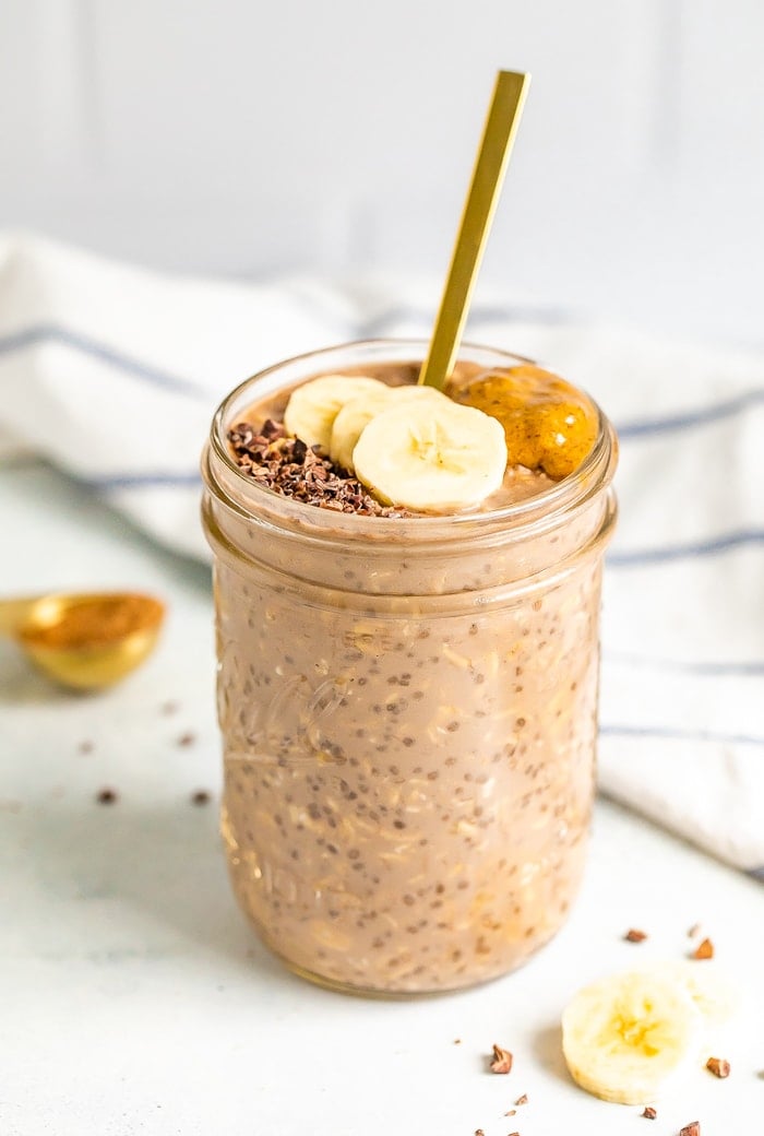A mason jar with chocolate banana overnight oats with a gold spoon handling sticking out. Oats are topped with banana slices, cacao nibs and almond butter.