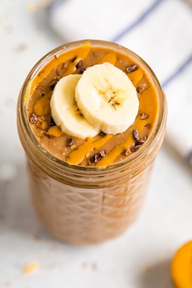 Jar filled with a chocolate smoothie topped with cacao nibs, banana slices and a peanut butter drizzle.