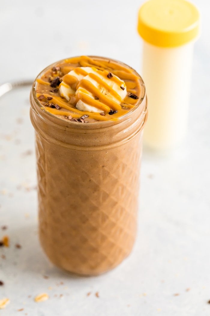 Jar filled with a chocolate smoothie topped with cacao nibs, banana slices and a peanut butter drizzle.