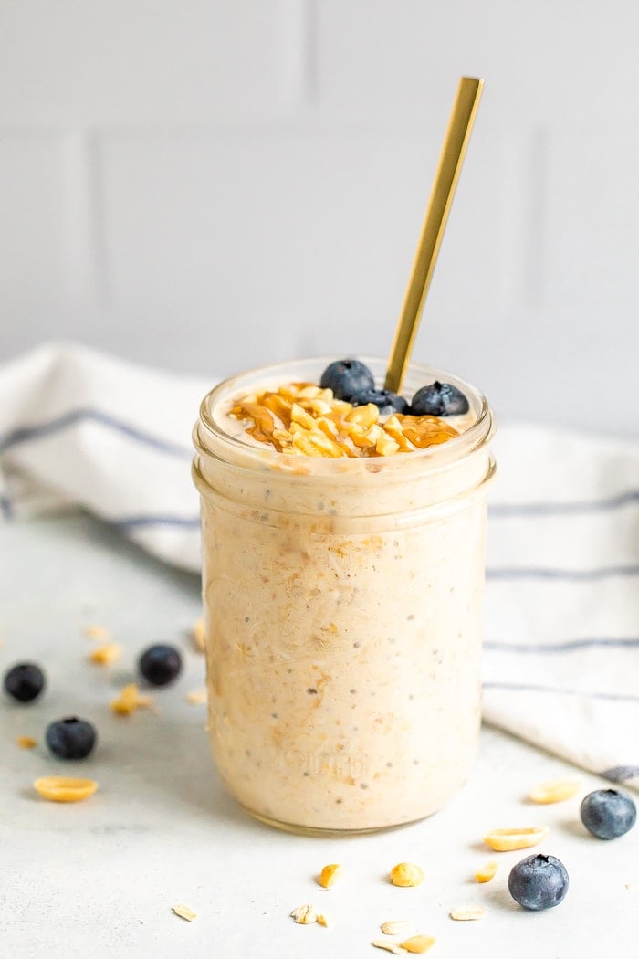 Peanut butter overnight oats in a mason jar with a gold spoon. Topped with blueberries, chopped peanuts and a peanut butter drizzle.