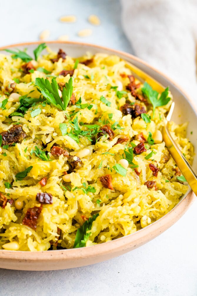 Bowl of pesto spaghetti squash salad topped with parsley and sun-dried tomatoes. Serving spoon is in the bowl.