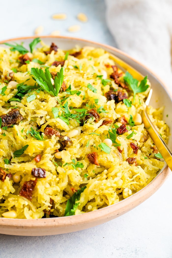 Bowl of pesto spaghetti squash salad topped with parsley and sun-dried tomatoes. Serving spoon is in the bowl.